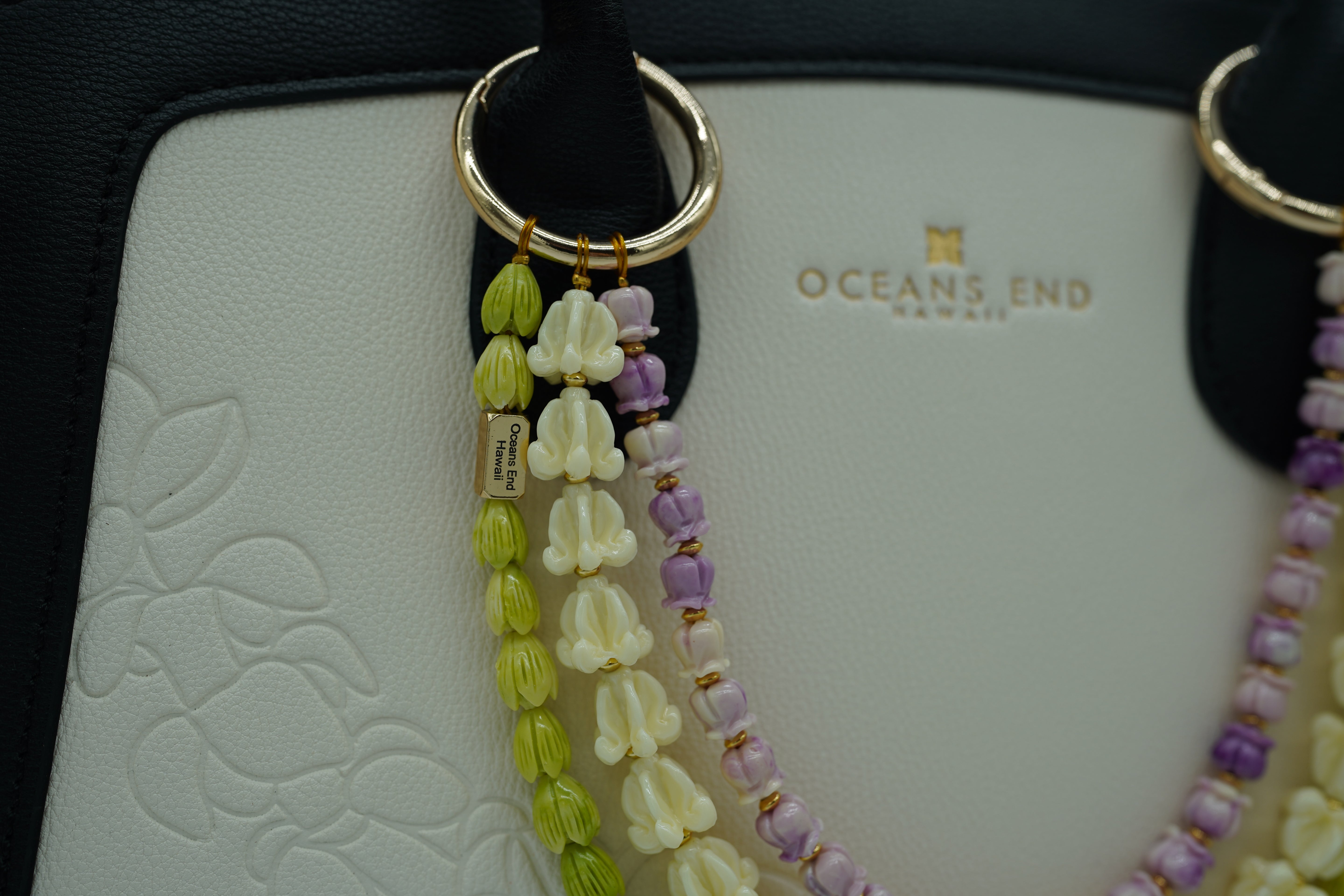 triple lei adornment featuring a green pikake flower lei strand on the bottom, a white crown flower lei strand in the middle and a purple crown flower lei strand on top with a small gold bar in the corner of the bottom lei showing our oceans end logo. lei adornment is attached to a bag using two gold rings. bag is sold separately.