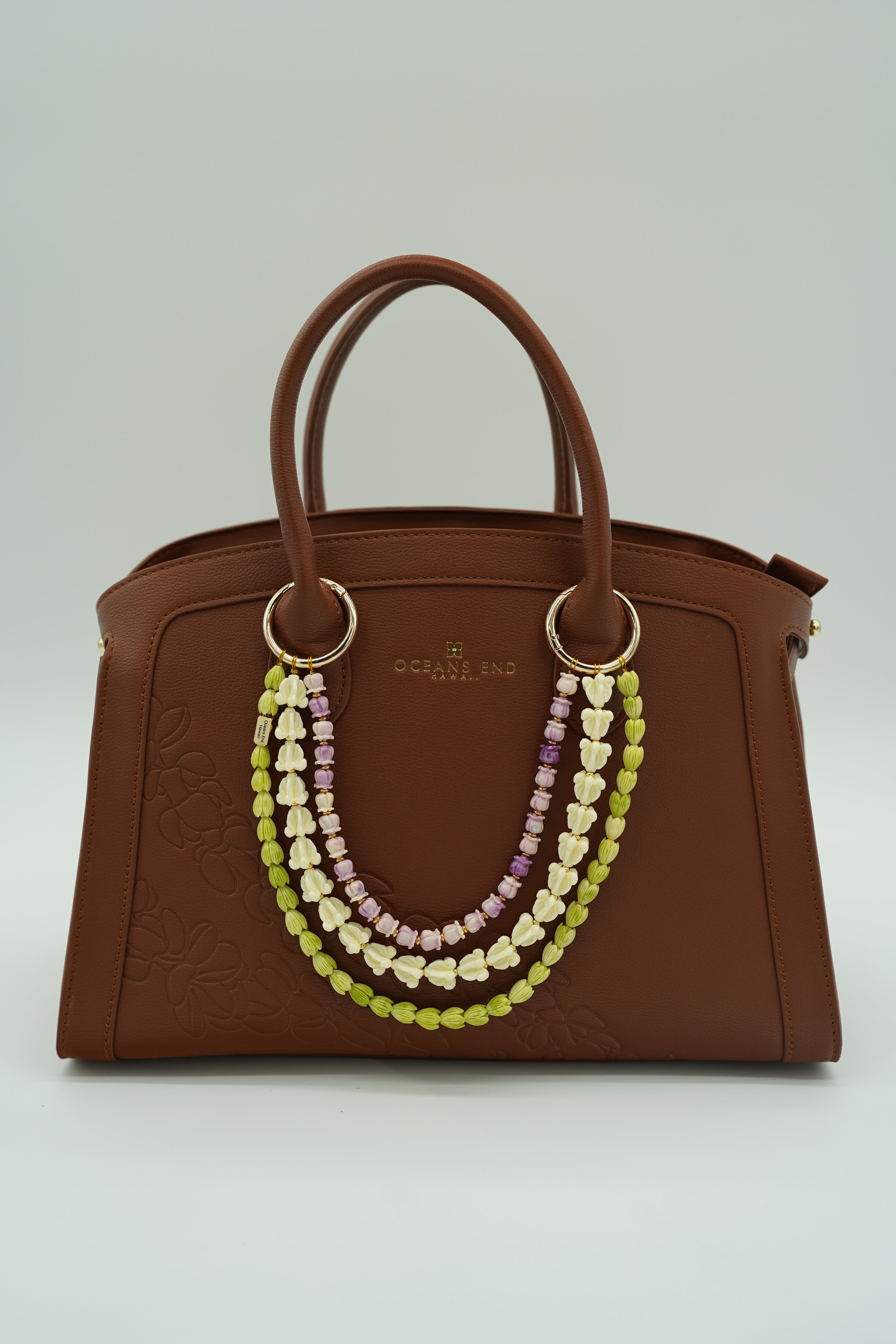 triple lei adornment featuring a green pikake flower lei strand on the bottom, a white crown flower lei strand in the middle and a purple crown flower lei strand on top with a small gold bar in the corner of the bottom lei showing our oceans end logo. lei adornment is attached to a bag using two gold rings. bag is sold separately.