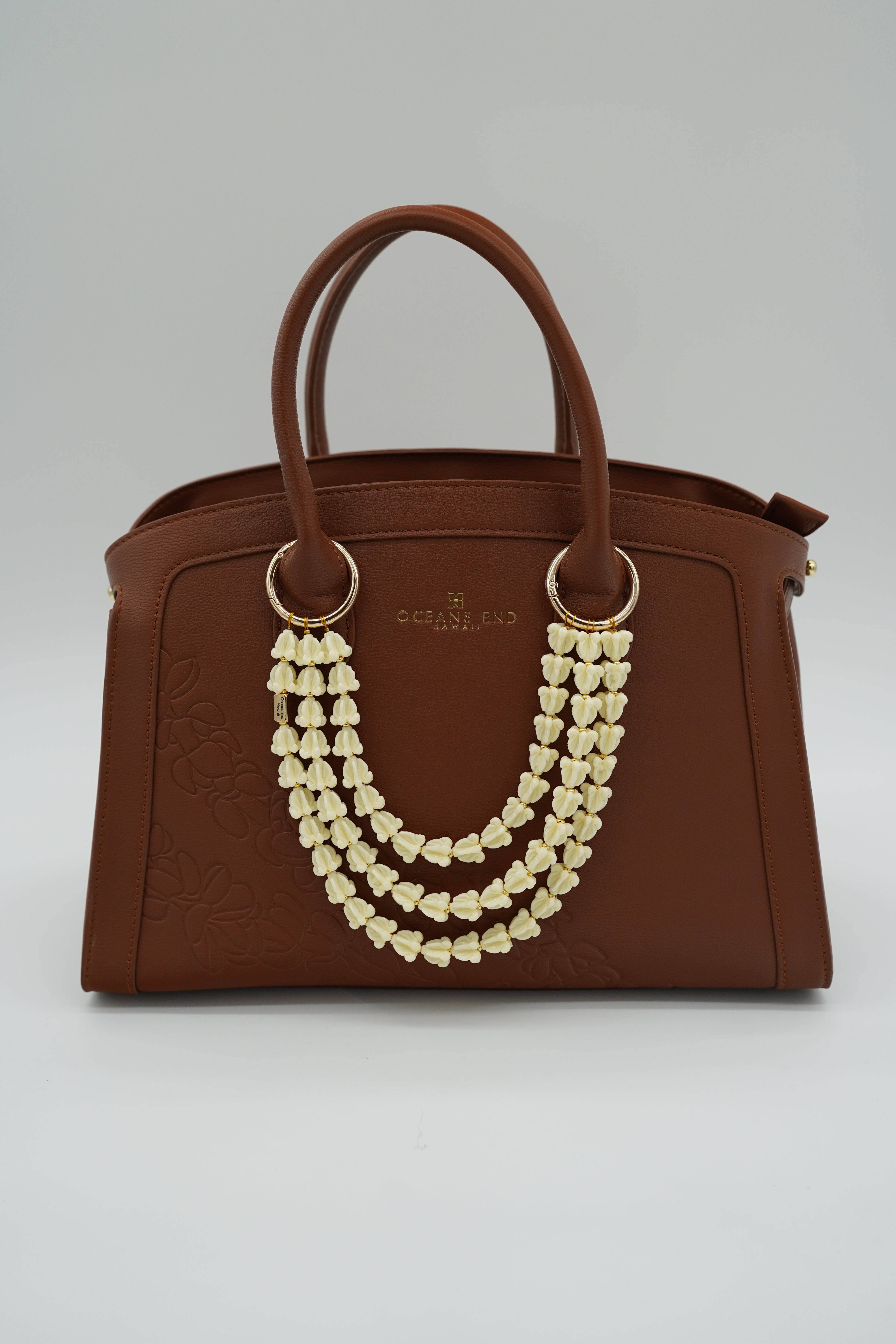 triple lei adornment featuring a white crow flower lei strand on the bottom, a white crown flower lei strand in the middle and a white crown flower lei strand on top with a small gold bar in the corner of the bottom lei showing our oceans end logo. lei adornment is attached to a bag using two gold rings. bag is sold separately.