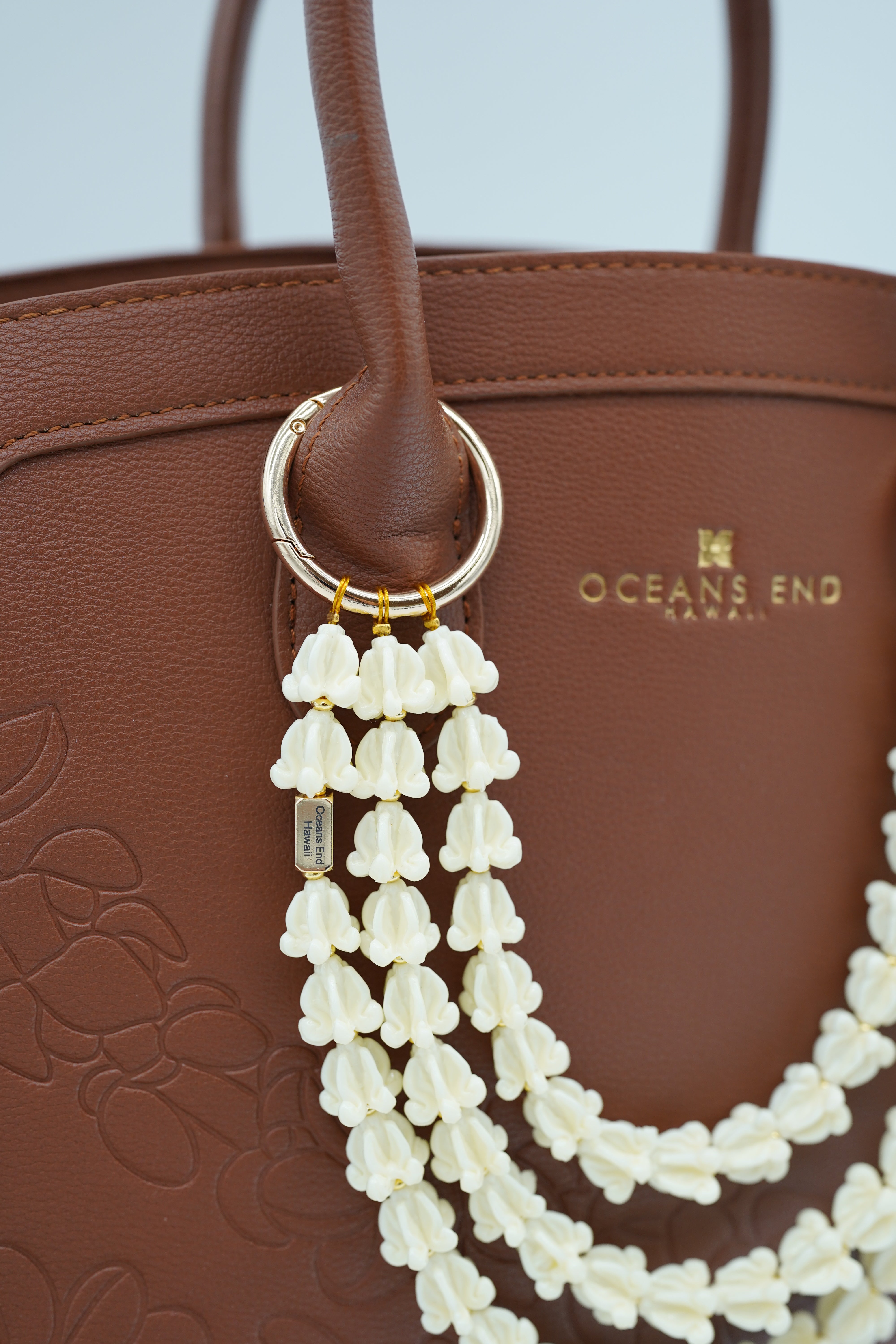 triple lei adornment featuring a white crow flower lei strand on the bottom, a white crown flower lei strand in the middle and a white crown flower lei strand on top with a small gold bar in the corner of the bottom lei showing our oceans end logo. lei adornment is attached to a bag using two gold rings. bag is sold separately.