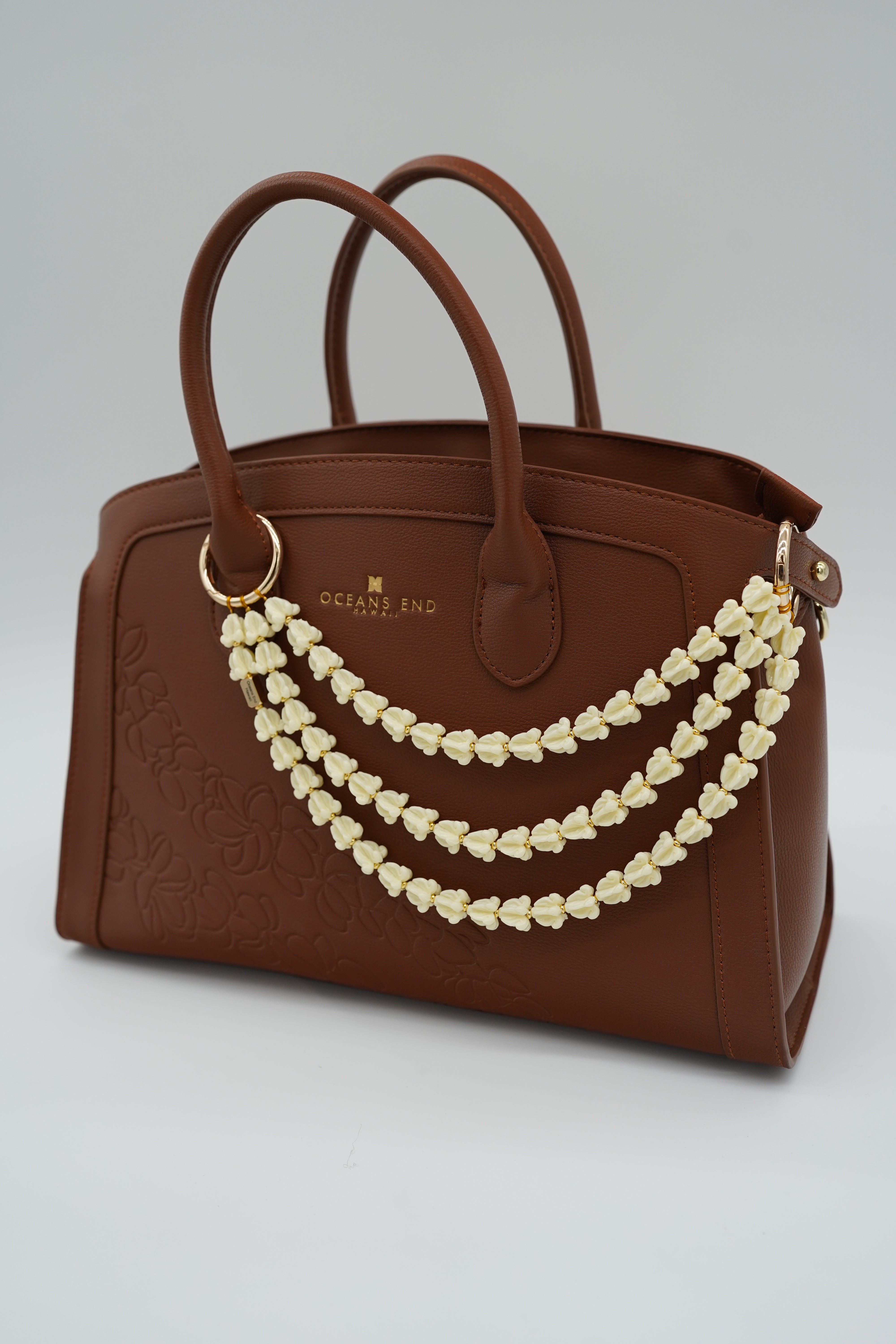 triple lei adornment featuring a white crow flower lei strand on the bottom, a white crown flower lei strand in the middle and a white crown flower lei strand on top with a small gold bar in the corner of the bottom lei showing our oceans end logo. lei adornment is attached to a bag using two gold rings. bag is sold separately.