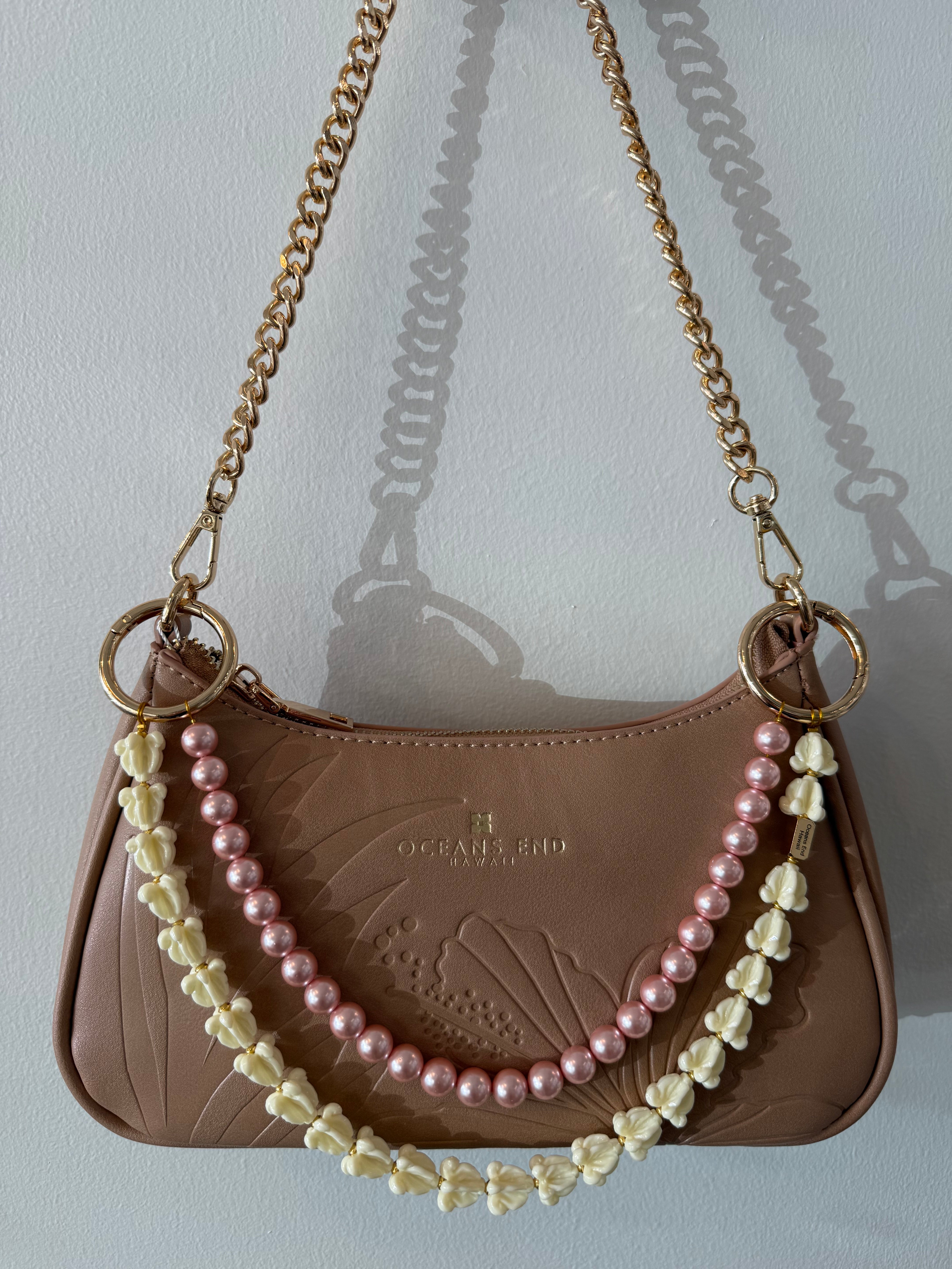double lei adornment with a white crown flower lei strand on the bottom and a pink pearl strand on top with a small gold bar in the corner of the bottom lei showing our oceans end logo. lei adornment is attached to a bag using gold loops. bag is sold separately.