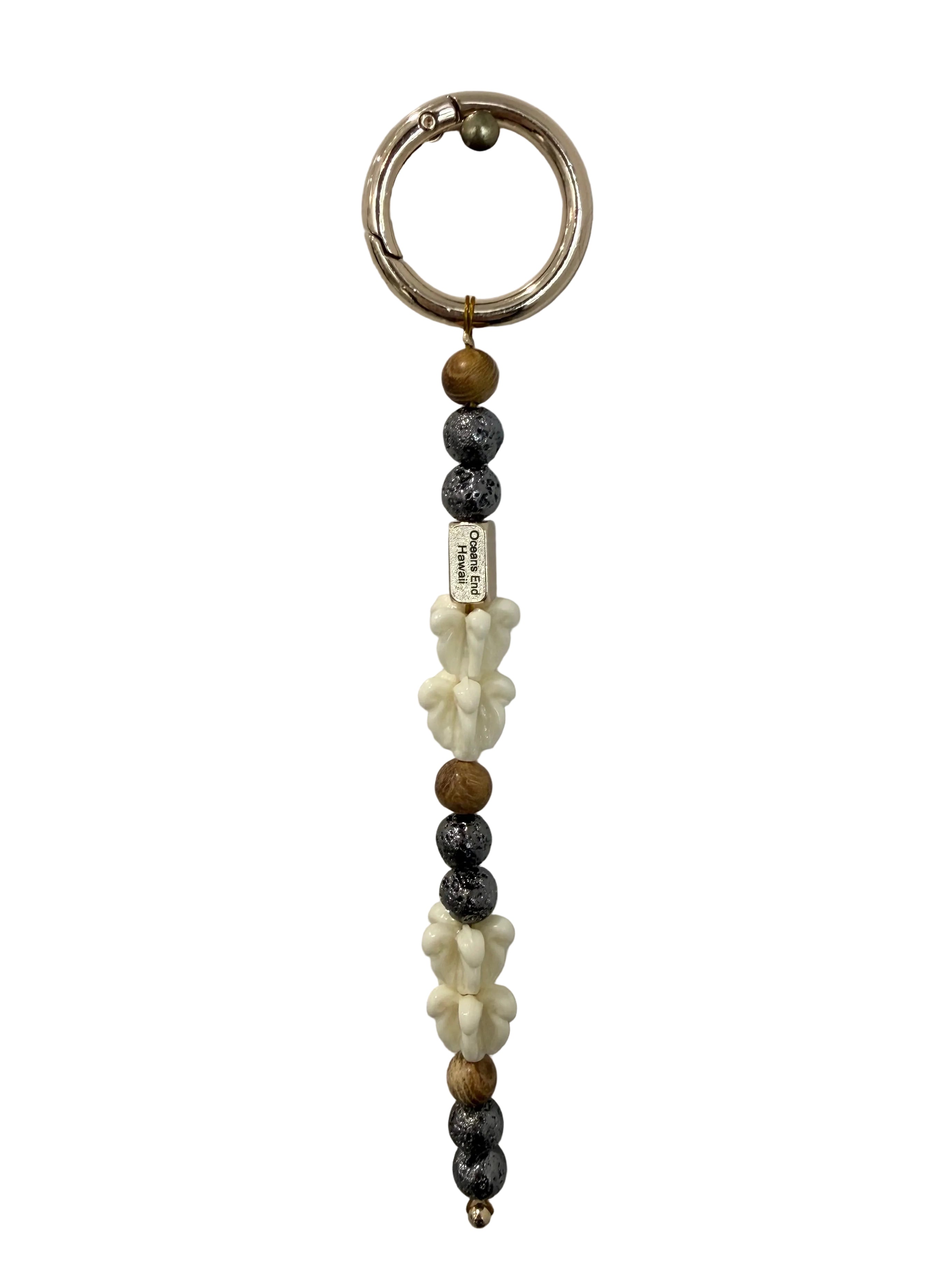 single lei keychain featuring white crown flower, wood and lava beads with a small gold bar showing our oceans end logo and a gold attachment hoop on top