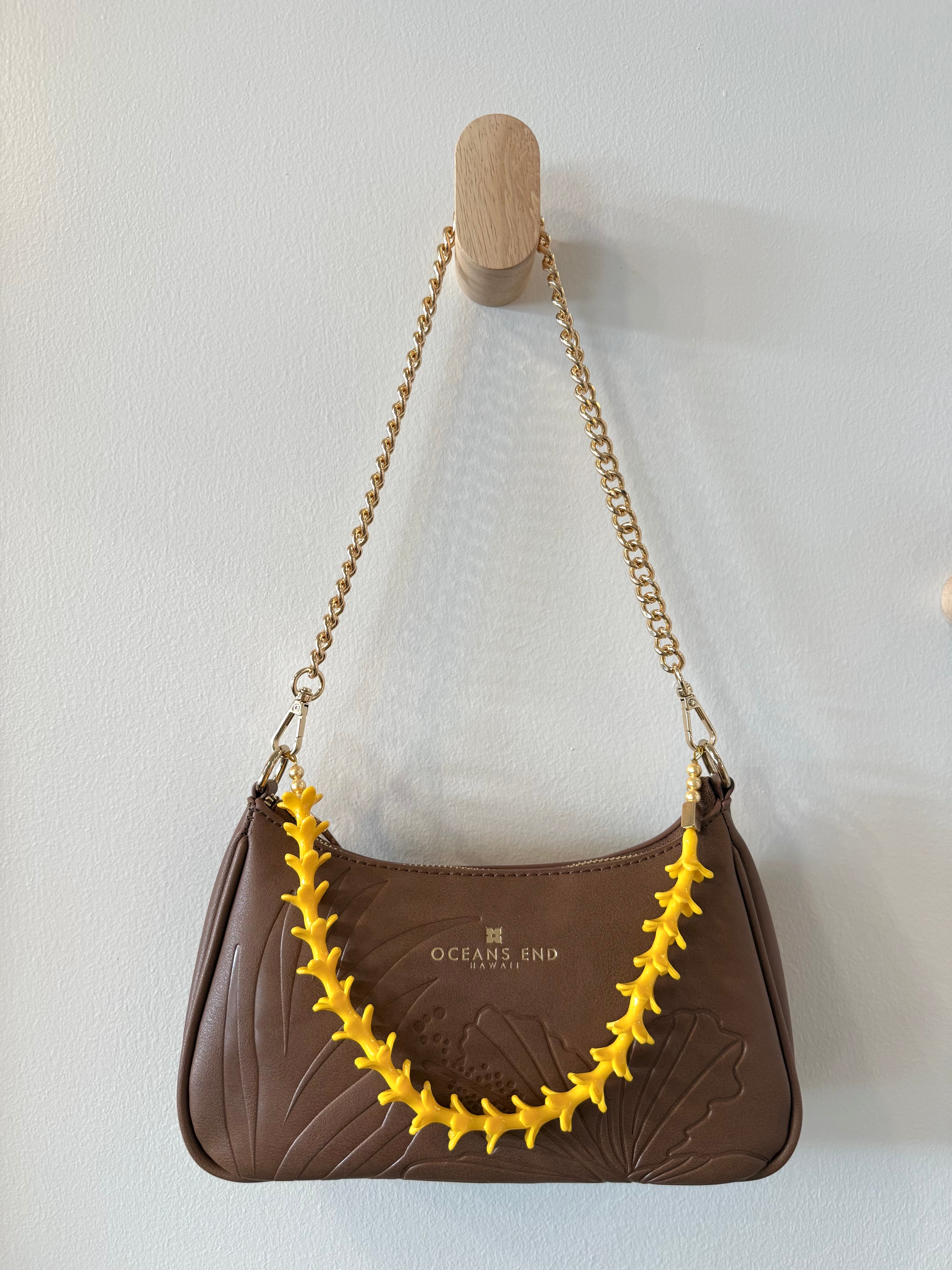 brown bag with hibiscus and leaf design on the front and our oceans end logo at the top center hanging from a gold chain shoulder strap with gold hardware featuring a yellow puakenikeni lei adornment attached.