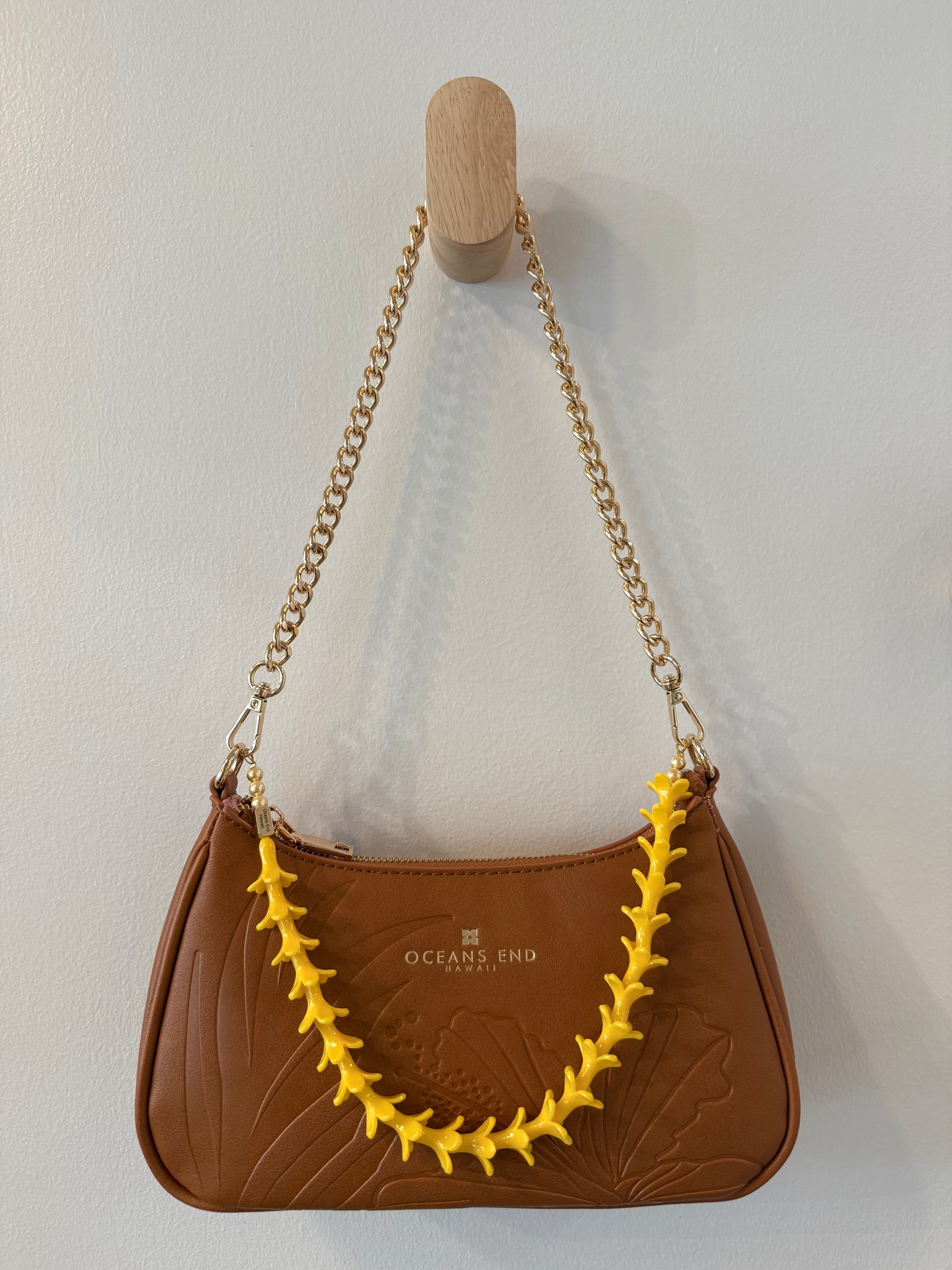 brown bag with hibiscus and leaf design on the front and our oceans end logo at the top center hanging from a gold chain shoulder strap with gold hardware featuring a yellow puakenikeni lei adornment attached.