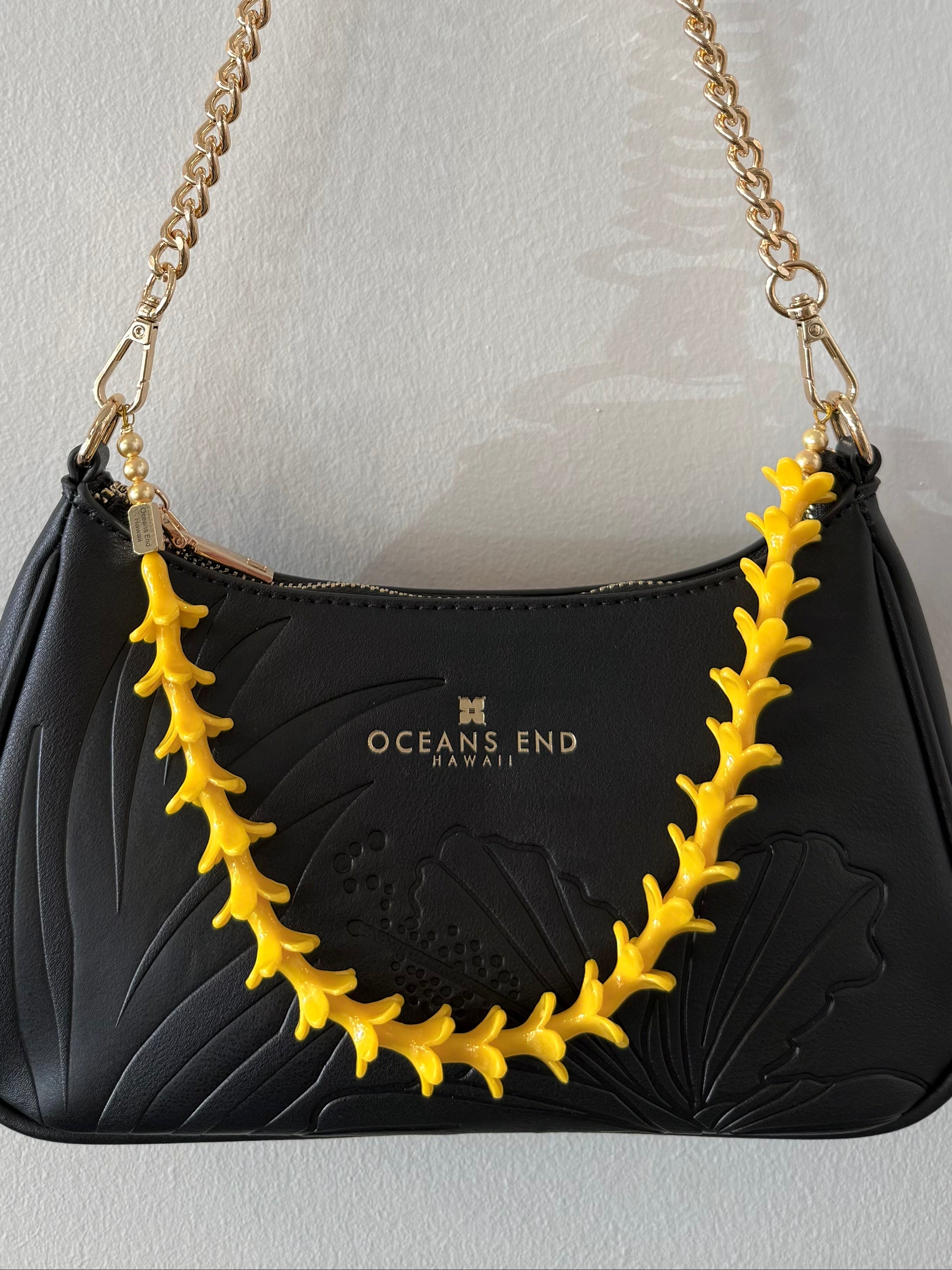 black bag with hibiscus and leaf design on the front and our oceans end logo at the top center hanging from a gold chain shoulder strap with gold hardware featuring a yellow puakenikeni lei adornment attached.