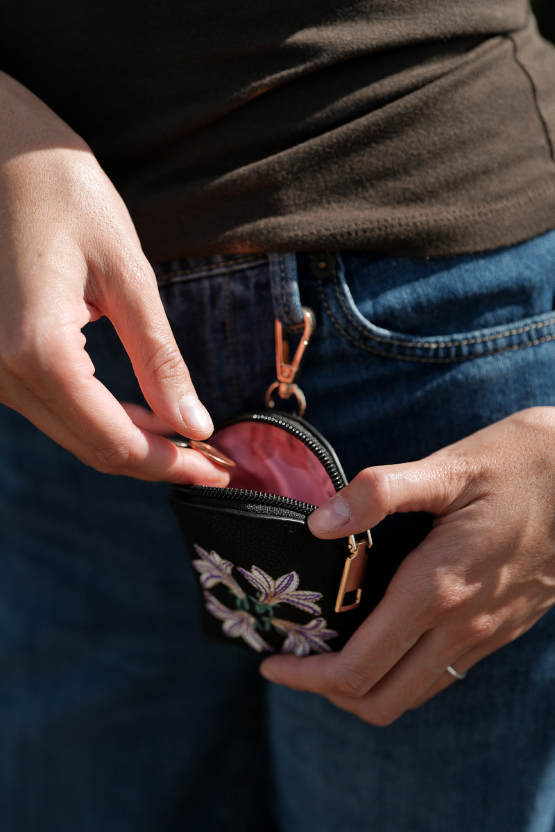 small pouch in the color black with naupaka design in the color white and purple and small green leafs in between featuring a gold zipper and gold claps at the top. displayed open on a jeans belt loop with coins put inside.