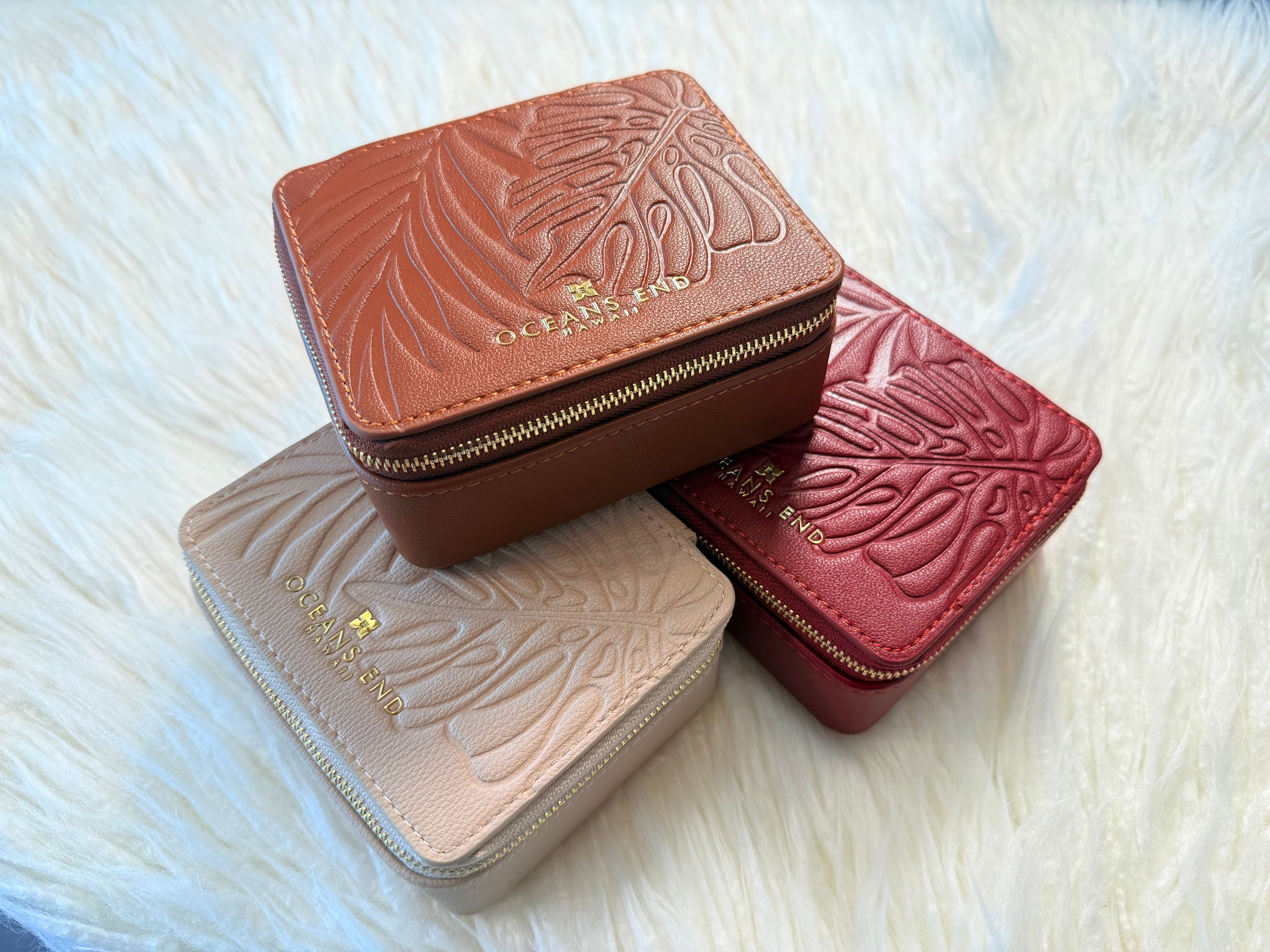 3 Compact jewelry cases stacked in the colors Rose, Taupe and Bourbon, gold oceans end logo with embossed monstera leaf detailing across exterior and a gold zip closure.