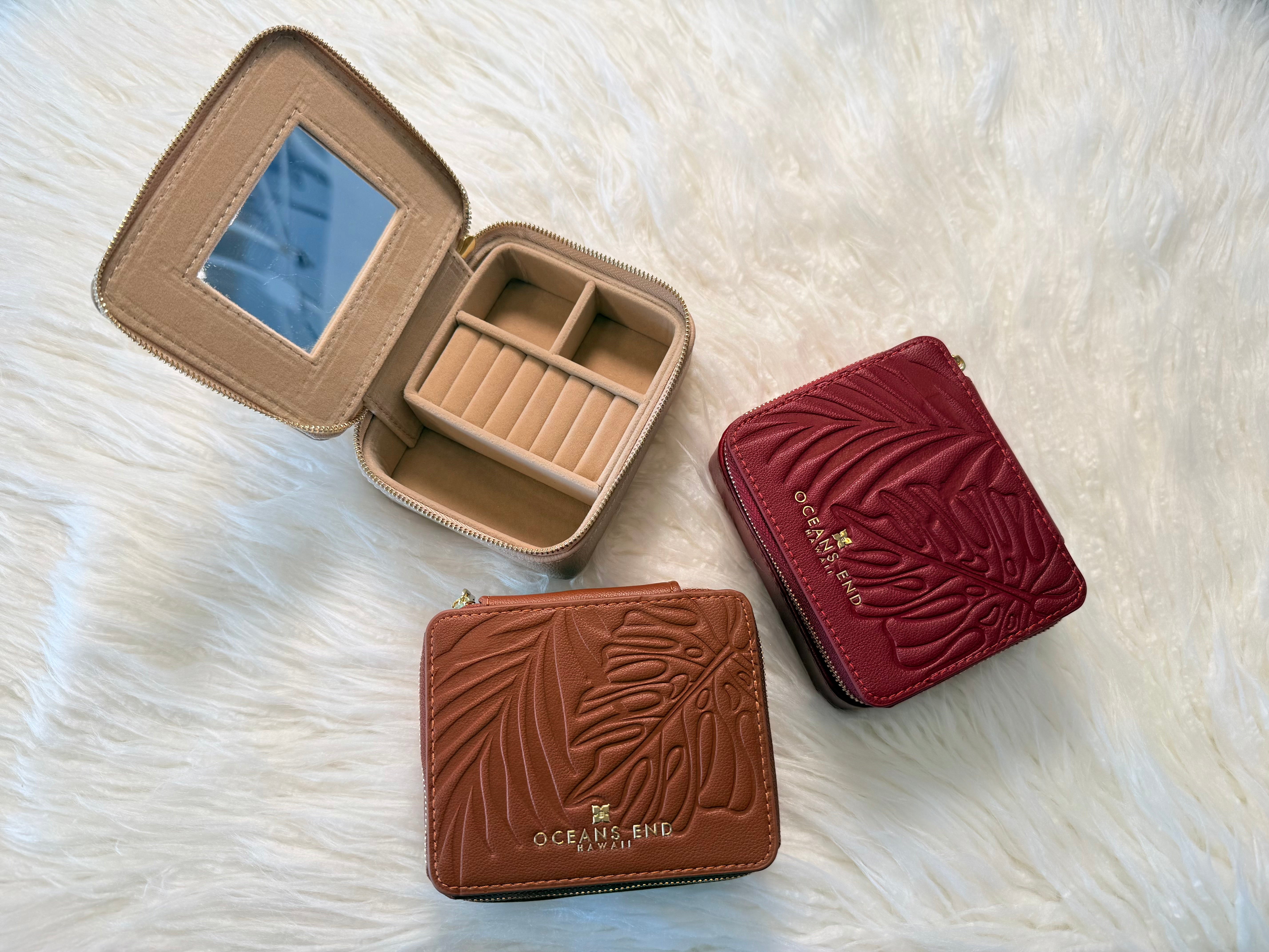 3 Compact jewelry cases in the colors Rose, Taupe and Bourbon, gold oceans end logo with embossed monstera leaf detailing across exterior and a gold zip closure. One of the jewelry cases are open showing the inside featuring a built in mirror on the lid, ring rolls, and 3 open compartments.